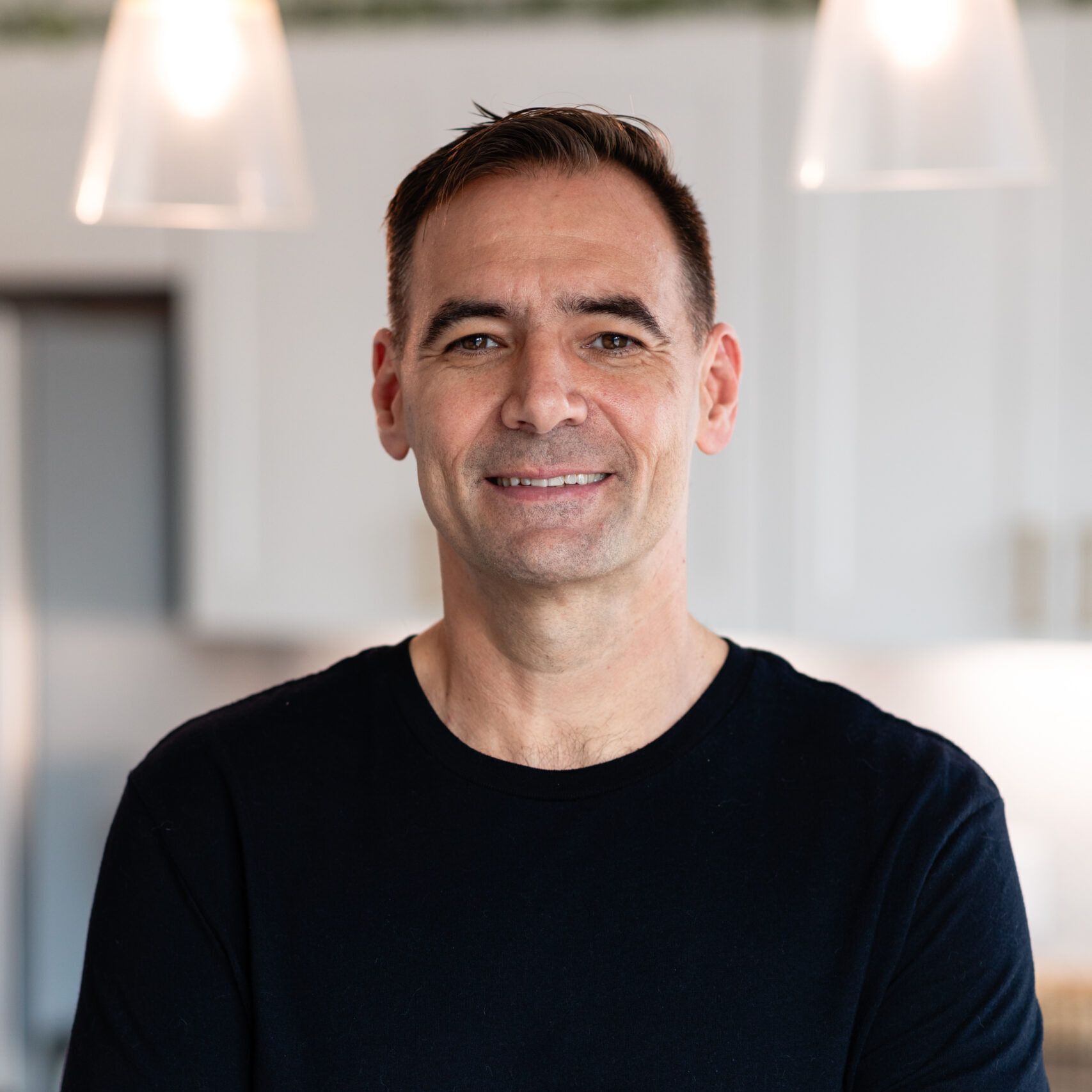 A man in a black shirt is smiling for the camera in a kitchen.