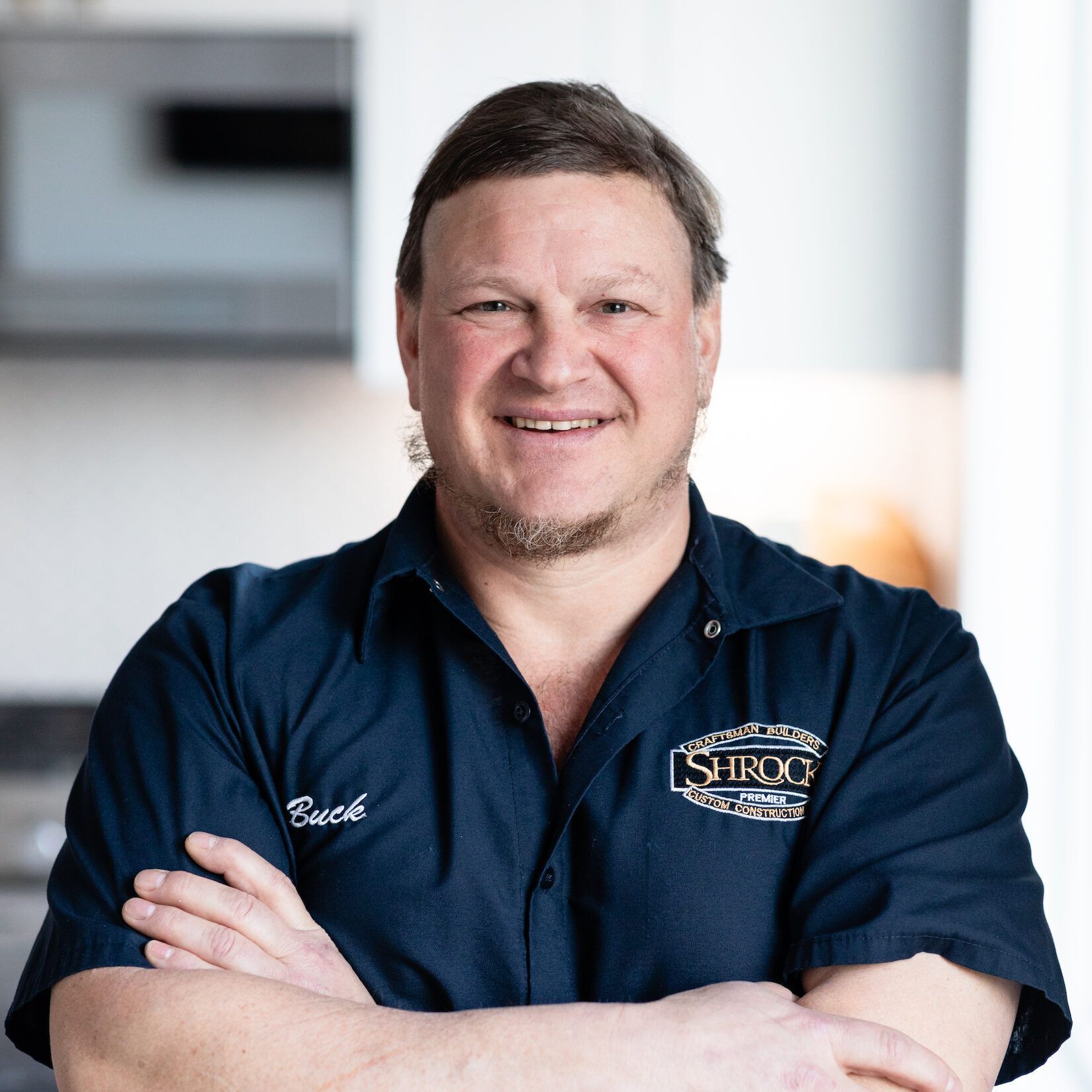 A man wearing a blue shirt with a Shrock logo on it is smiling with his arms crossed.