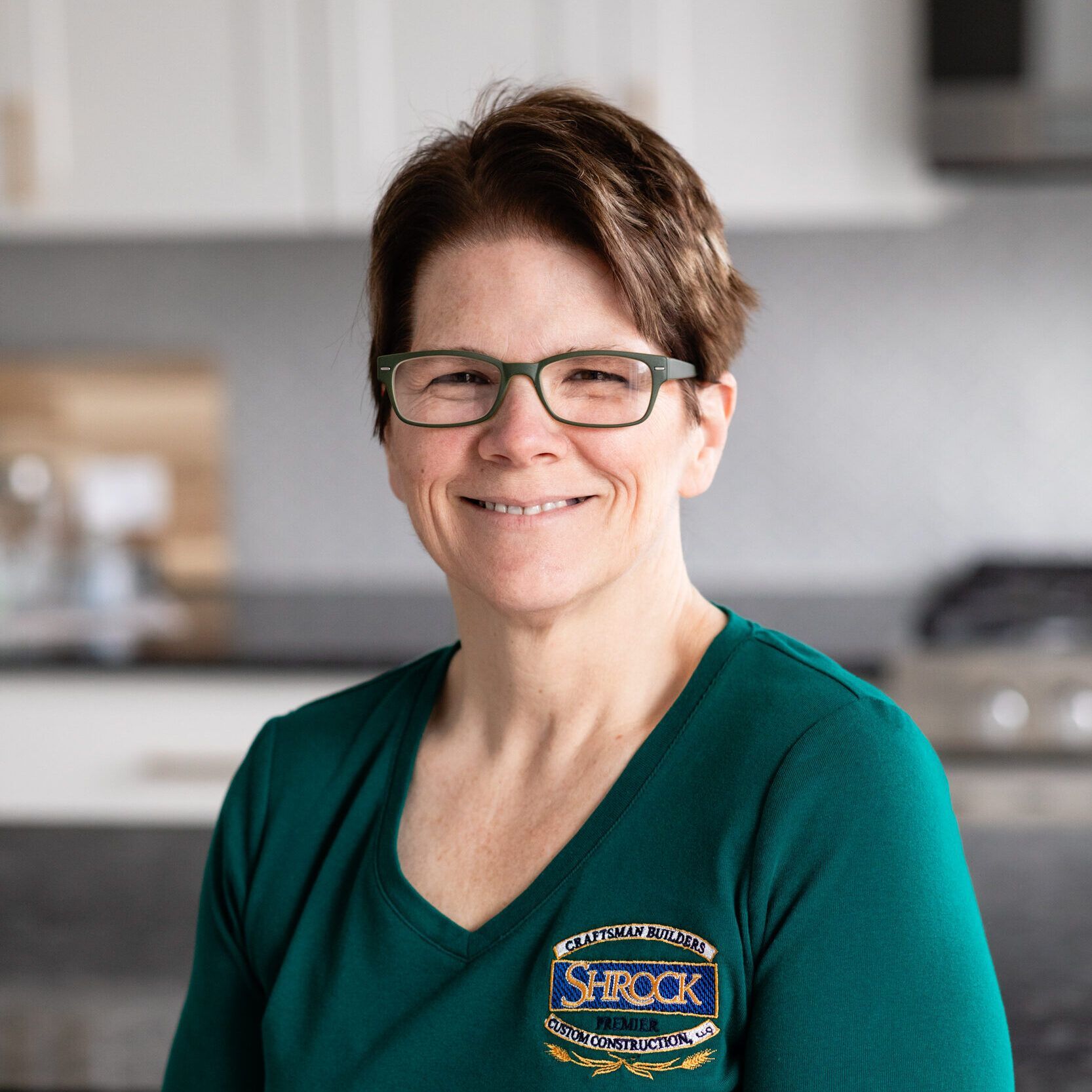 A woman wearing glasses and a green shirt is smiling in a kitchen.