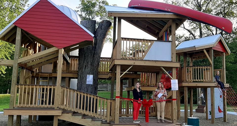 Two people are cutting a red ribbon in front of a tree house.