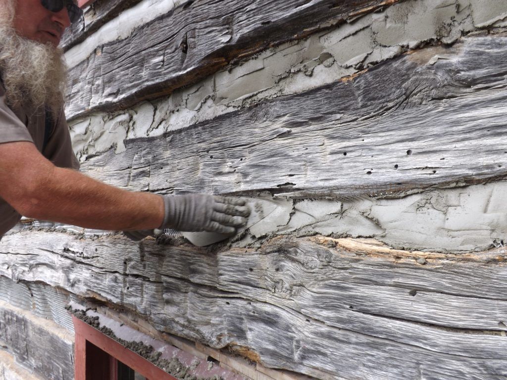 A man with a beard is working on the side of a log cabin.