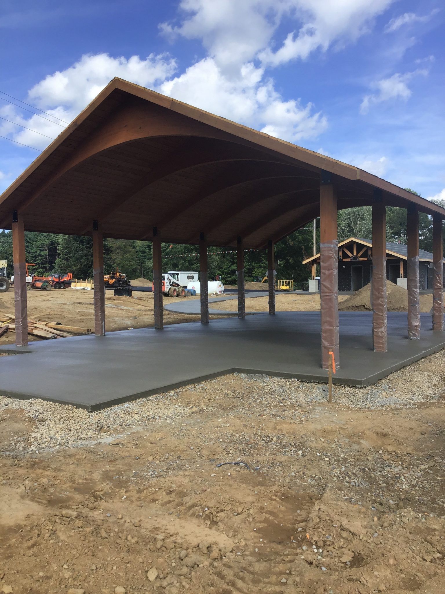 A wooden pavilion with a concrete floor underneath it is being built in a dirt field.