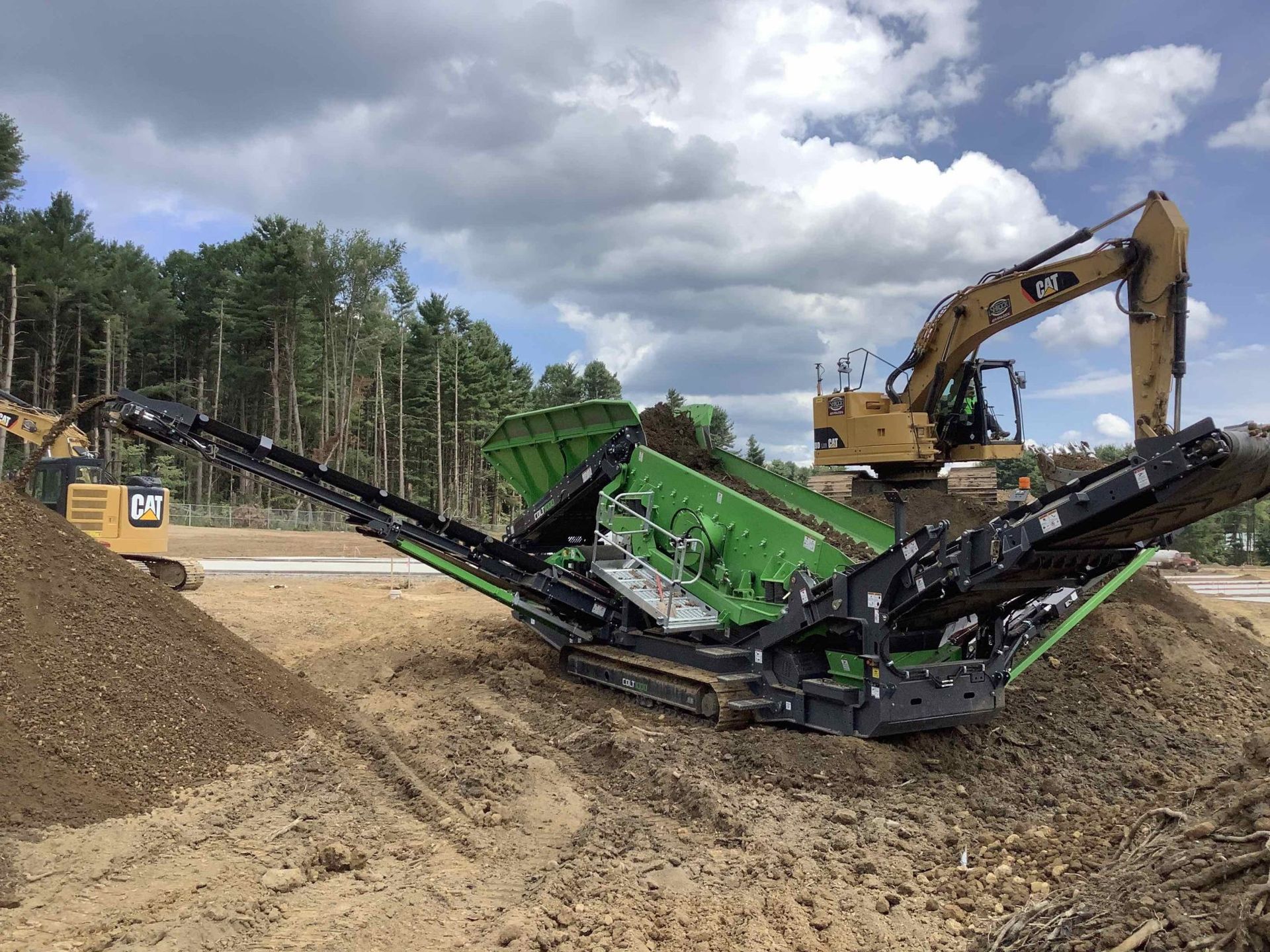 A green and black machine is sitting on top of a pile of dirt next to an excavator.