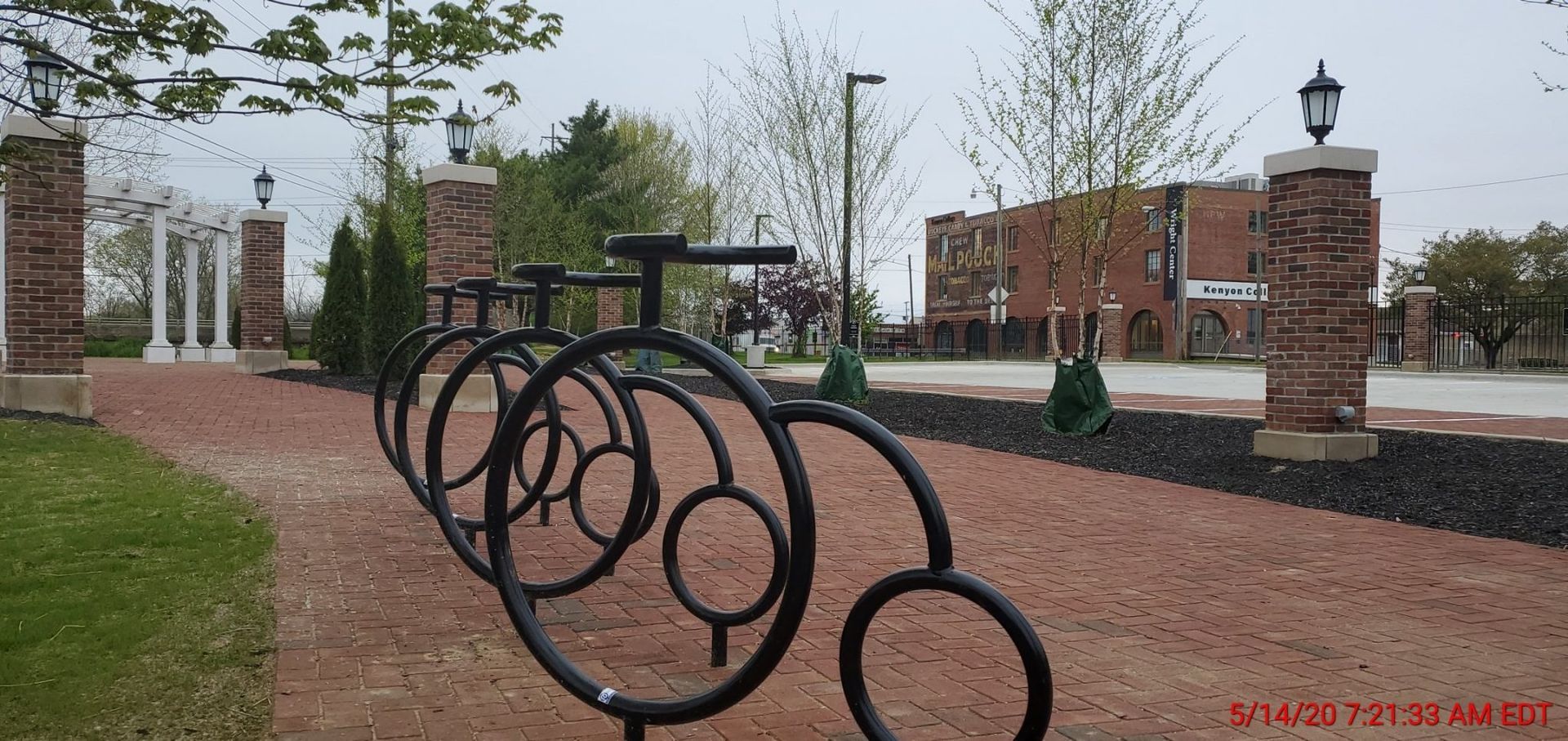 A row of bicycle racks in a park with a brick building in the background.