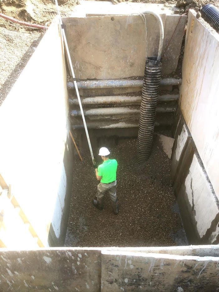 A man in a green shirt and hard hat is kneeling in a hole filled with gravel.