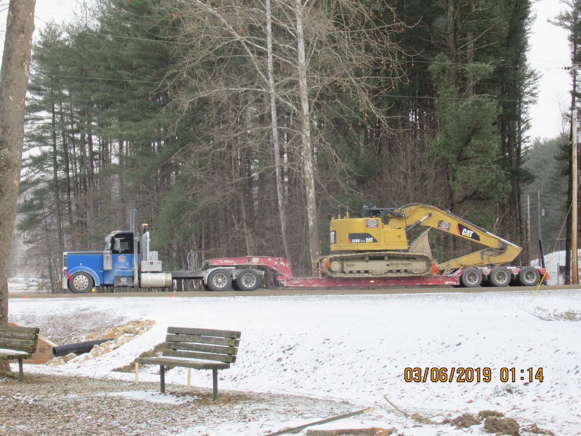 A blue truck is carrying a yellow excavator down a snowy road.