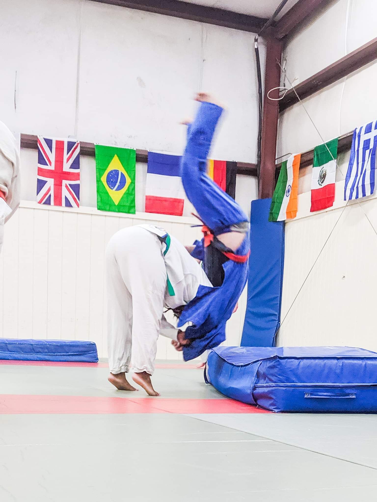 Two people are practicing judo in a gym with flags hanging on the wall.