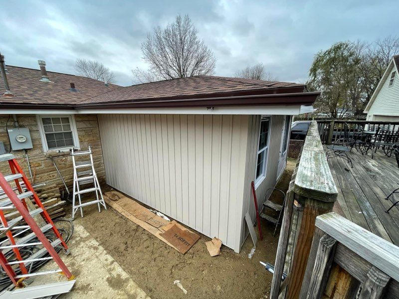 House exterior with vertical beige siding, roof overhang, ladders, and a narrow dirt yard between fences