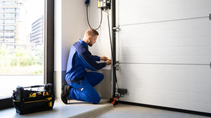 A man is kneeling down in a garage fixing a garage door.