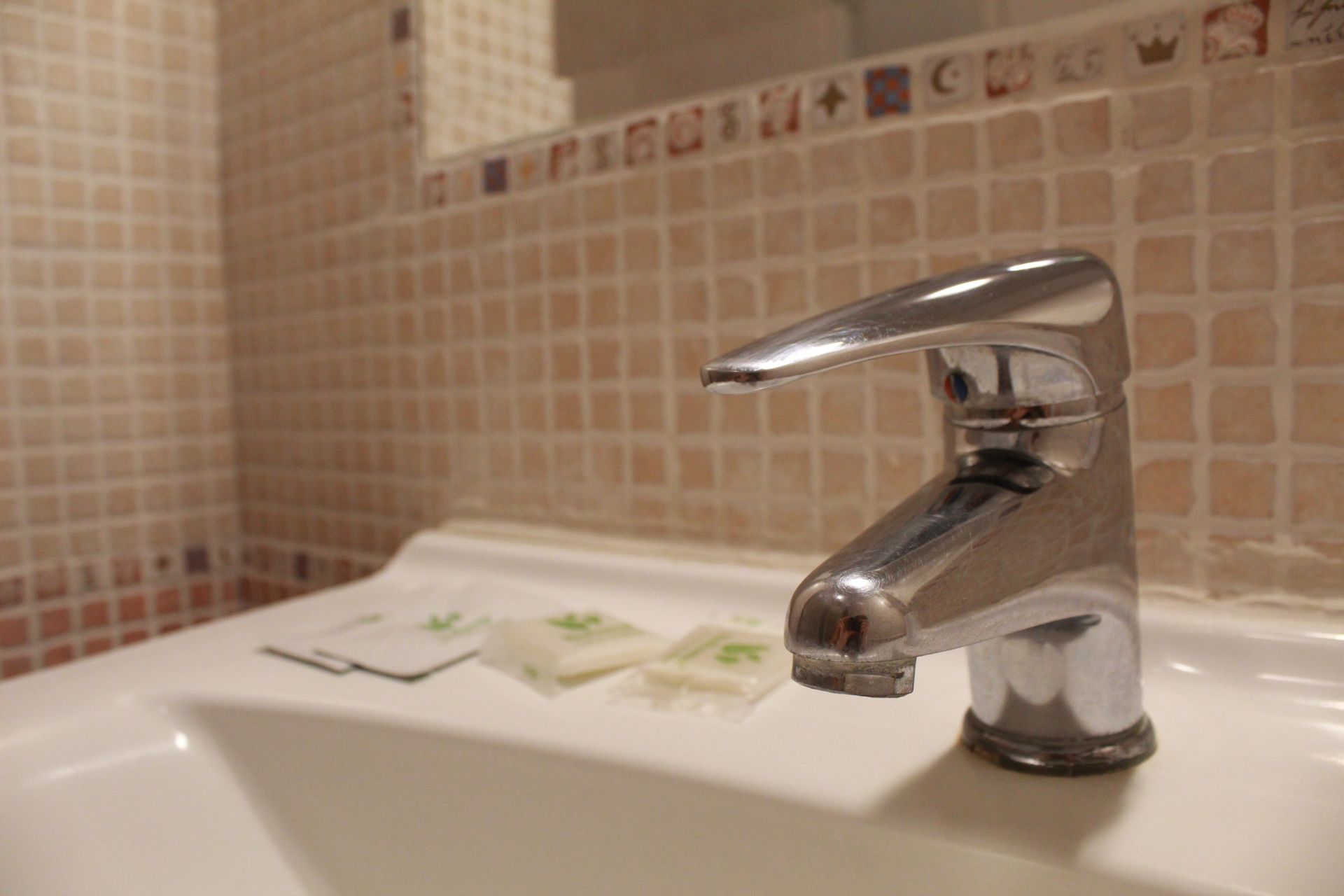 Rubinetto cromato lucido sopra un lavandino bianco con bustine di sapone davanti a una parete piastrellata.