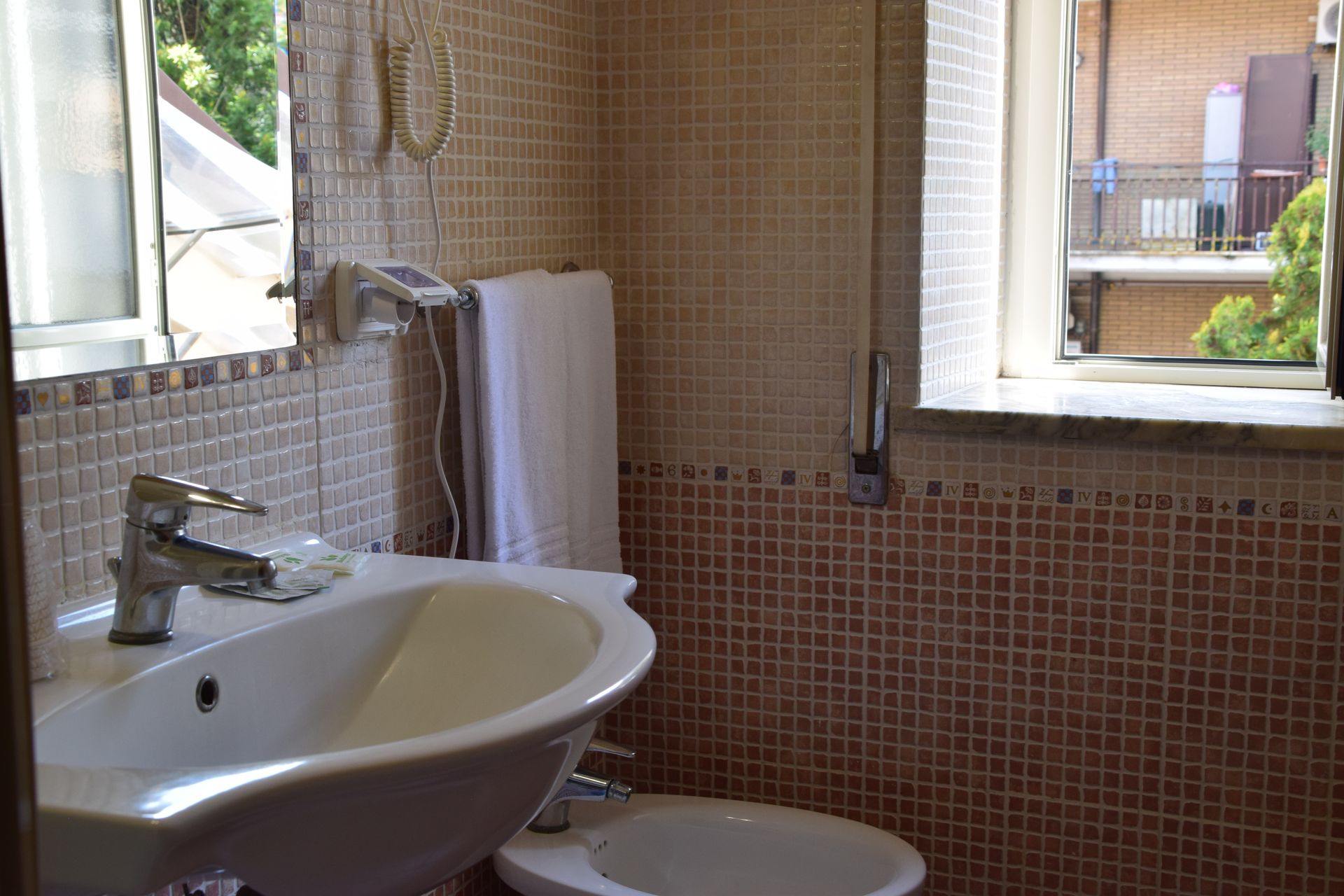 Piccolo bagno con pareti piastrellate. Lavandino, WC, finestra con vista, specchio e asciugamano.