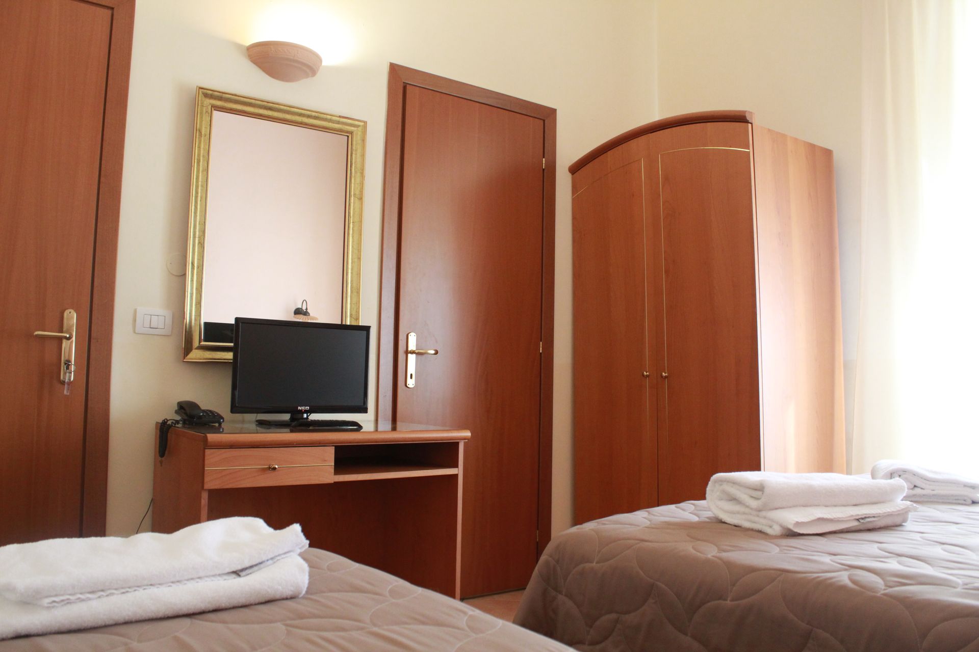 Camera d'albergo con due letti, scrivania con TV, specchio, porta in legno e armadio.