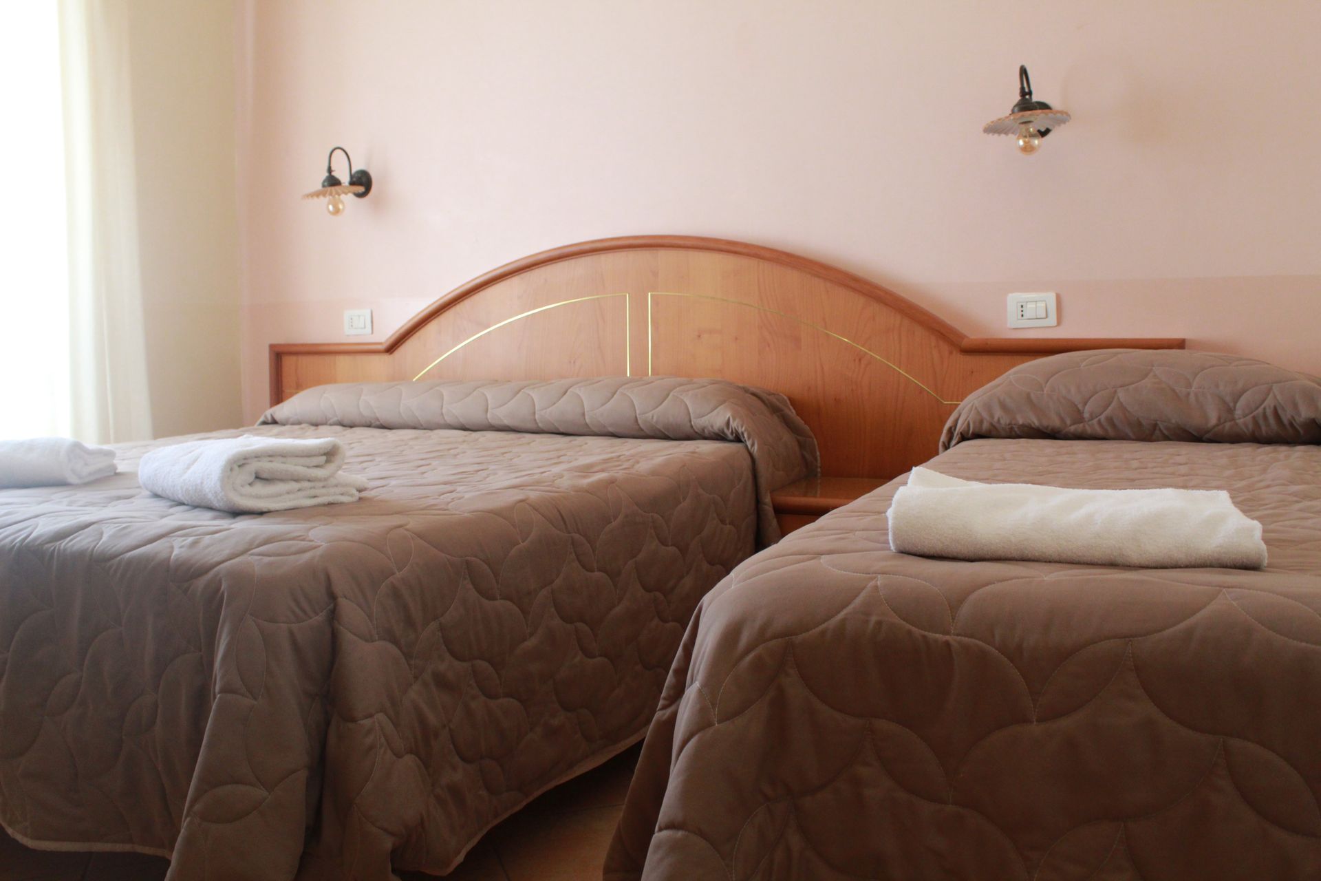 Due letti con piumoni marroni, asciugamani bianchi e lampade su una parete rosa.