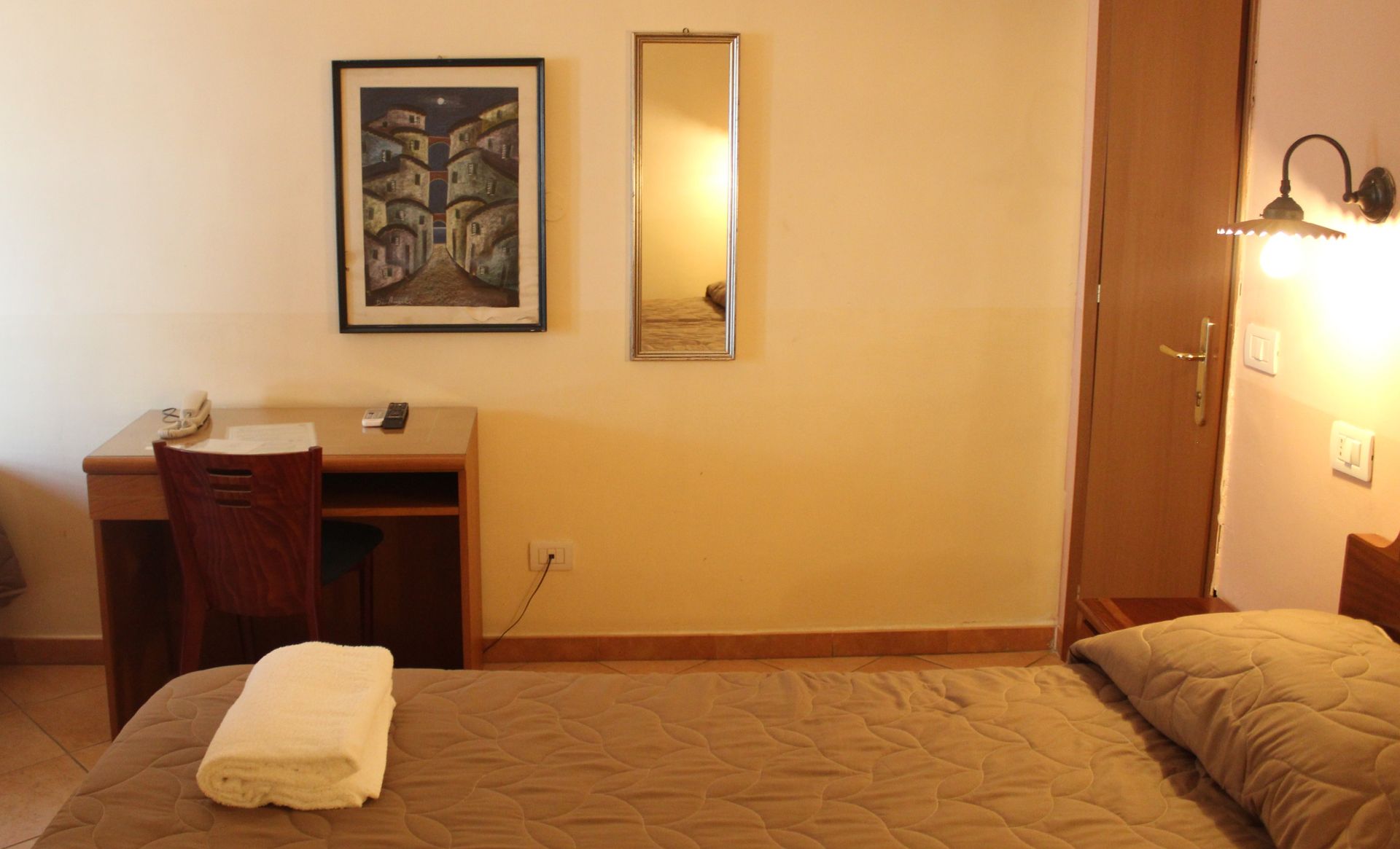 Camera d'albergo con letto, scrivania, opere d'arte, specchio e armadio. Pareti color crema, lenzuola marroni e un asciugamano.