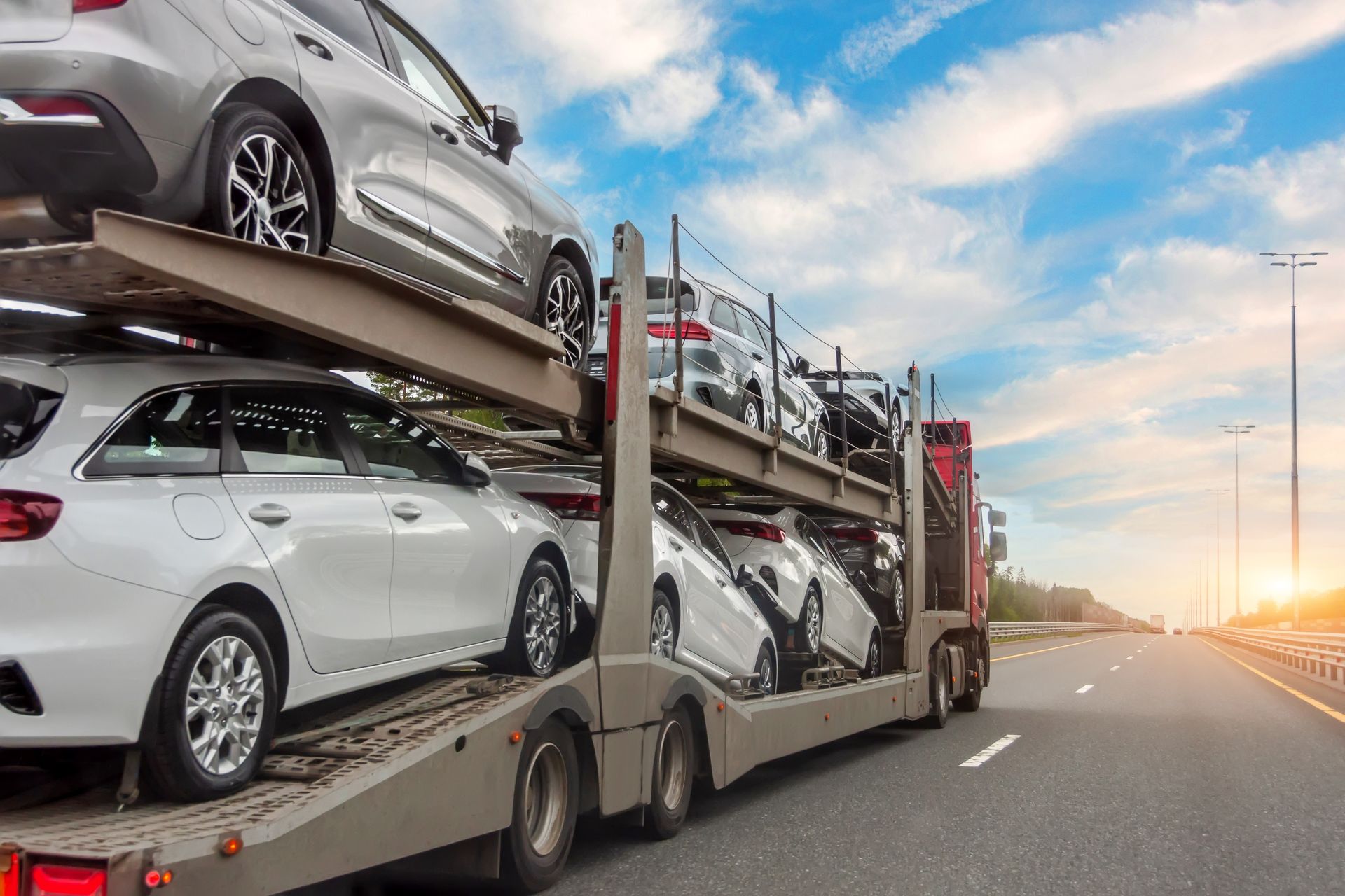 Car carrier truck transporting multiple new white and silver cars on a highway, sunny sky.