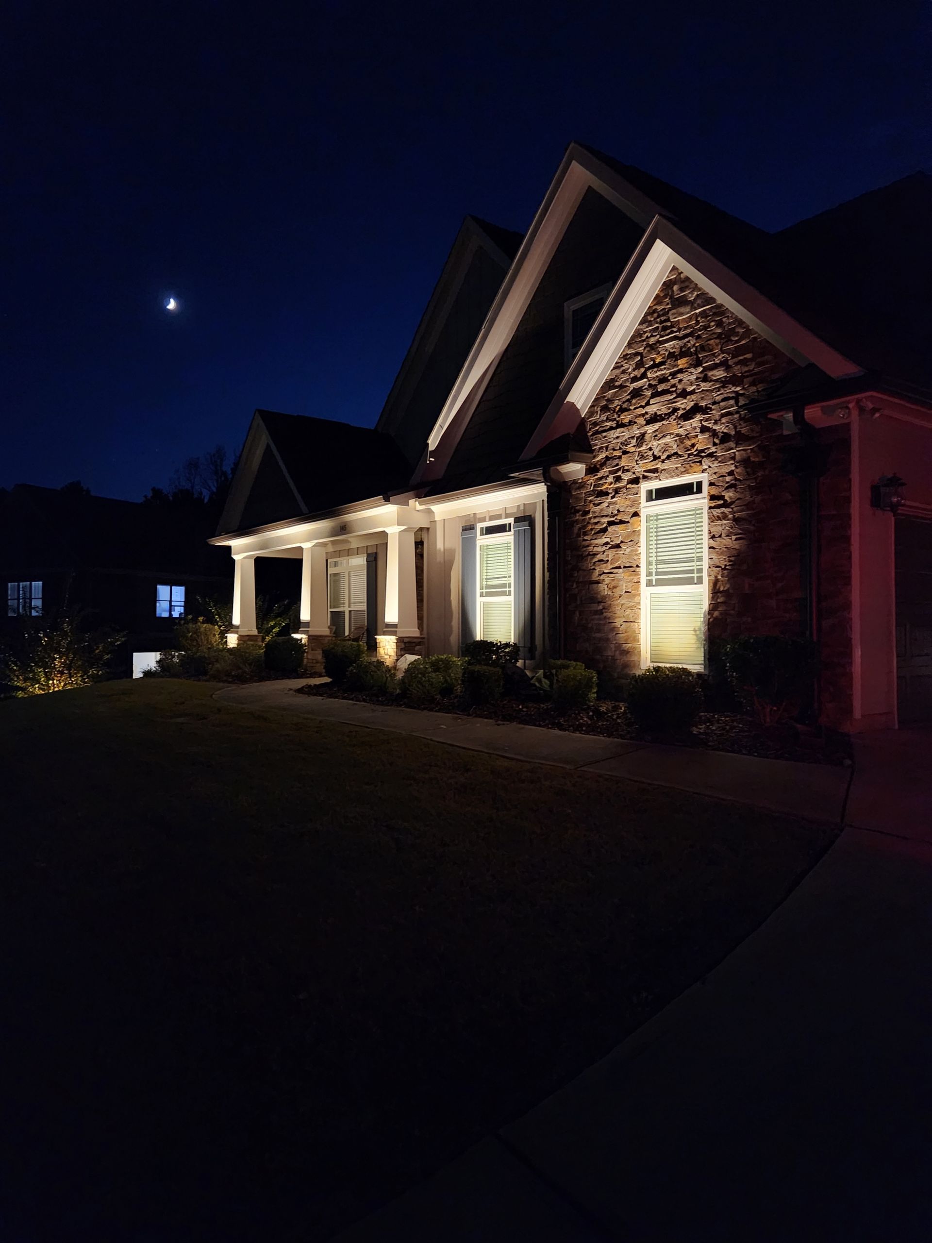 Lit-up house at night, stone facade and white trim, under a dark blue sky with a crescent moon.