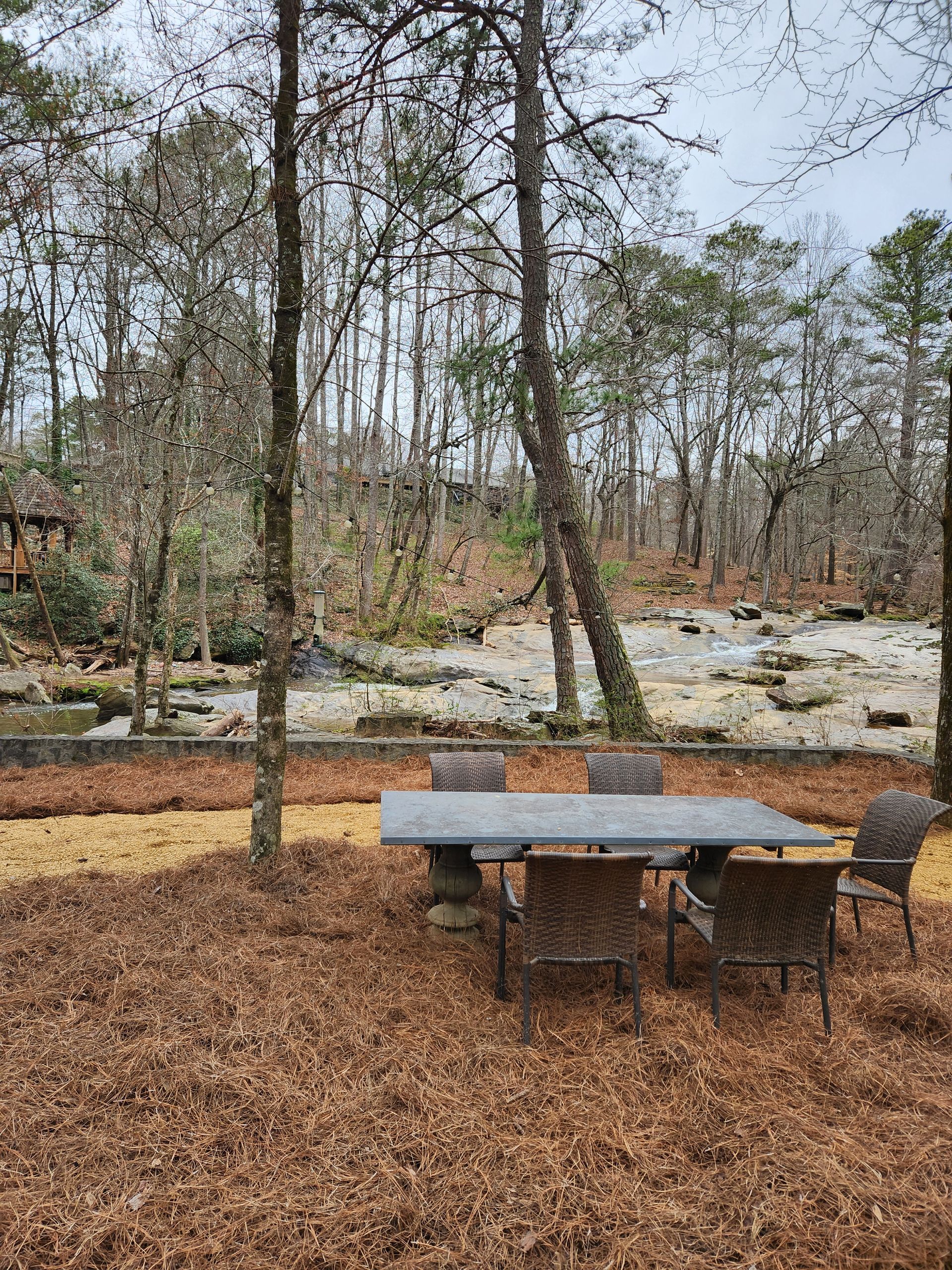 Outdoor dining set on brown mulch near a stream in a forest.