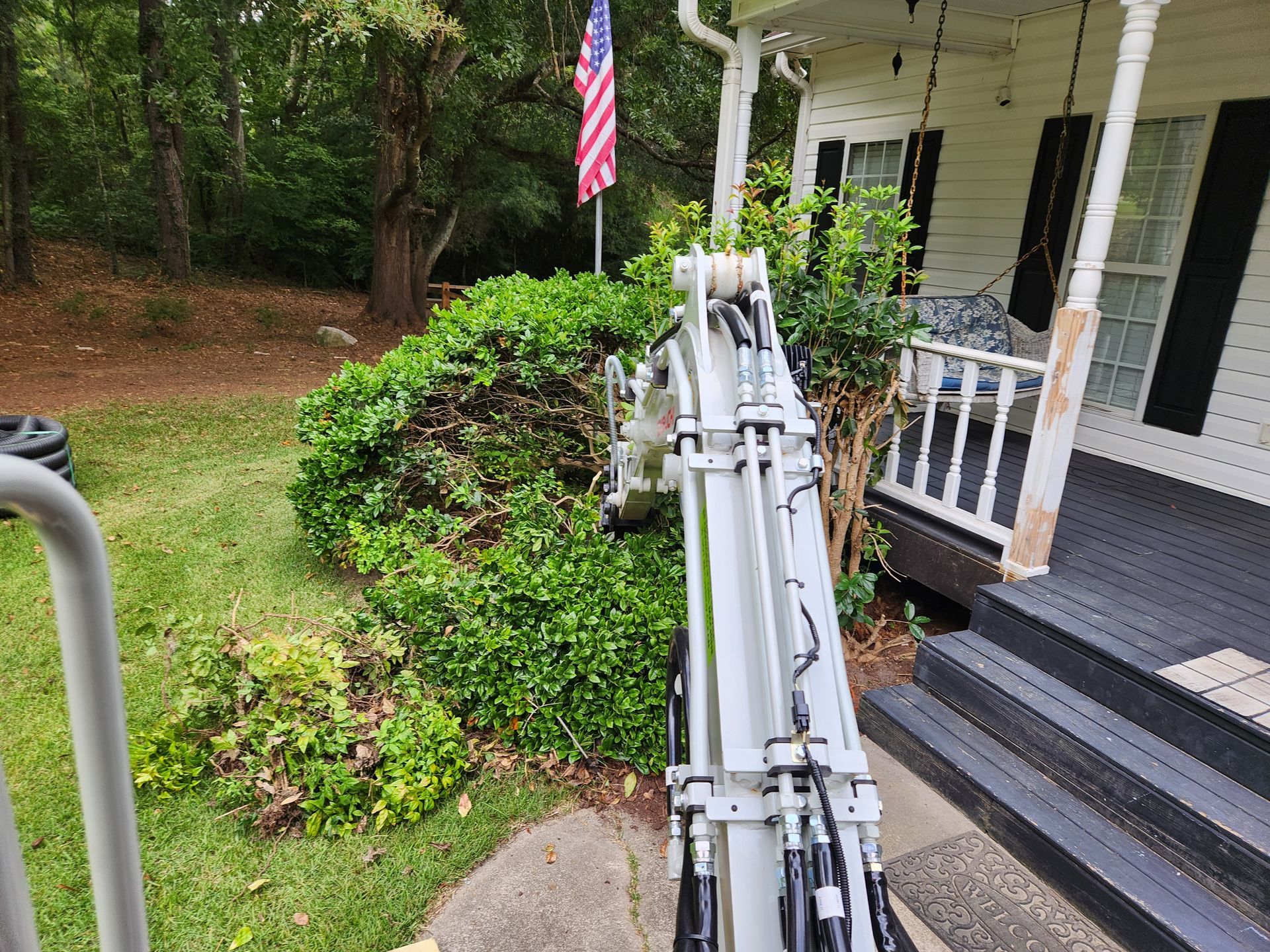 A white excavator is trimming a green bush next to a white house with a porch. An American flag is visible.