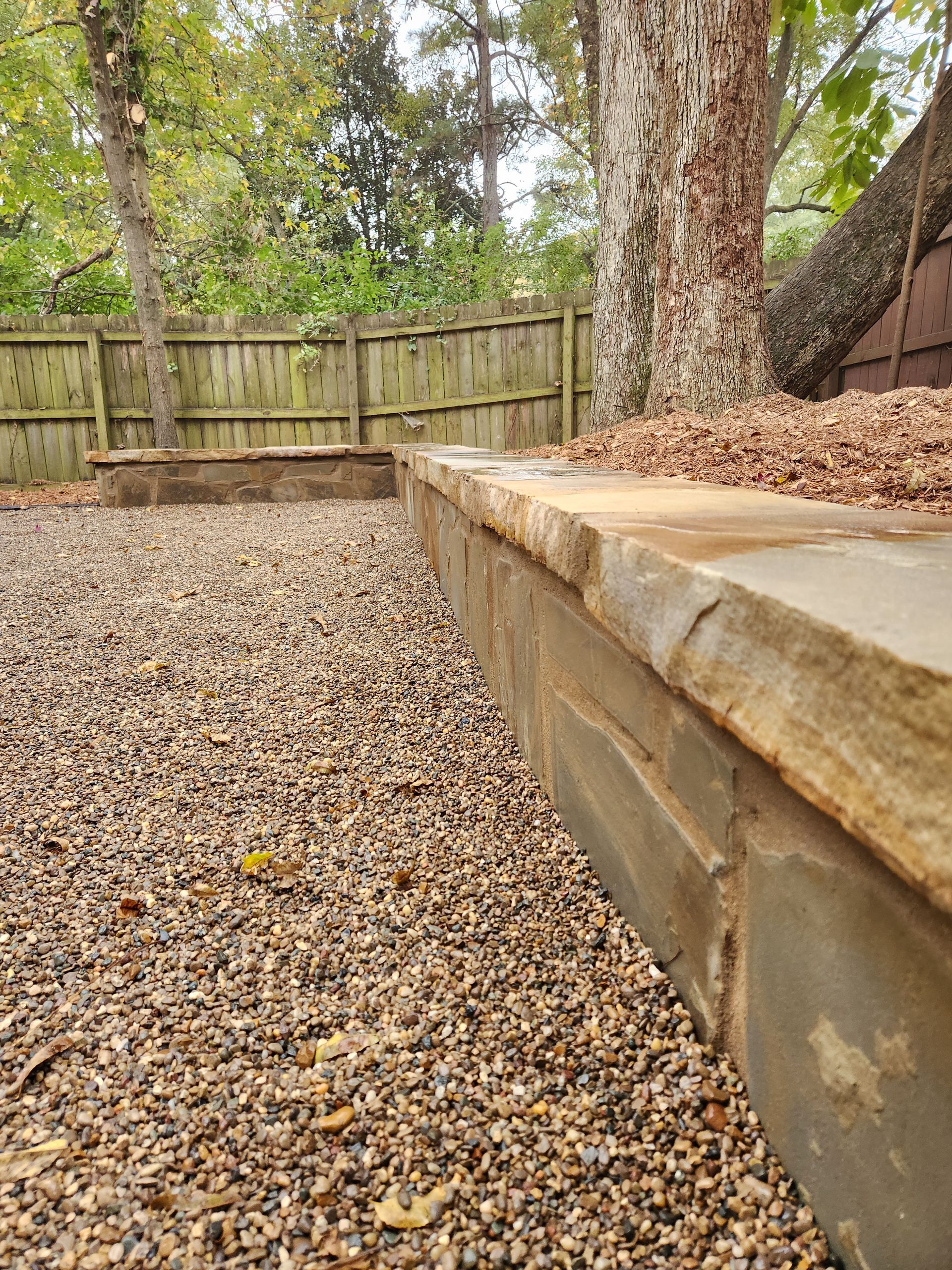 Stone retaining wall borders gravel pathway; wooden fence and trees in the background.