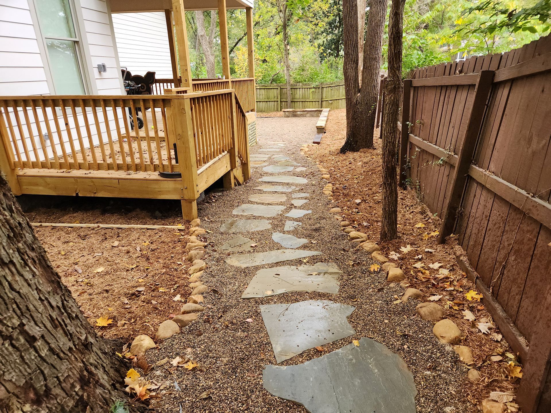 Stone pathway leads to a wooden porch, flanked by a wooden fence and trees. Brown leaves and gravel line the path.