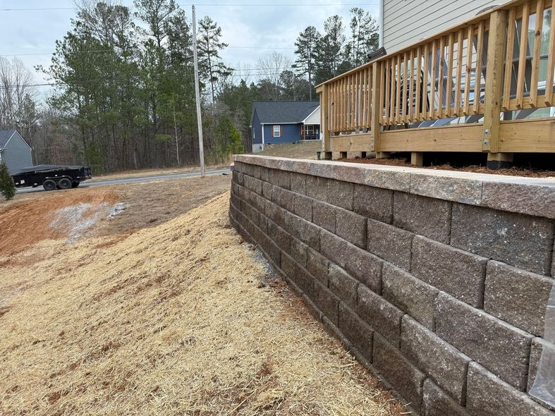 Stone retaining wall next to a wooden deck and a dirt/gravel slope. House in the background.