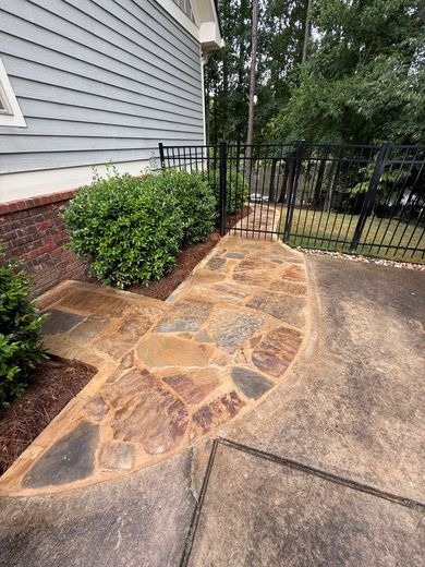 Stone pathway leading to a black gate, adjacent to a house and greenery.