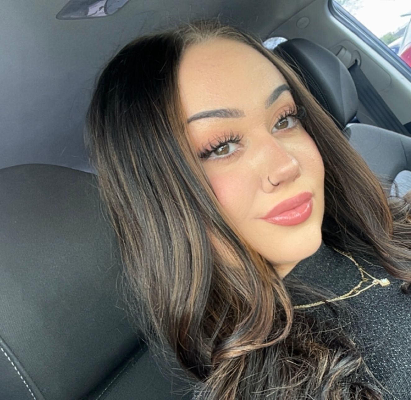 Woman with dark hair, nose ring, wearing a necklace, smiling in a car.