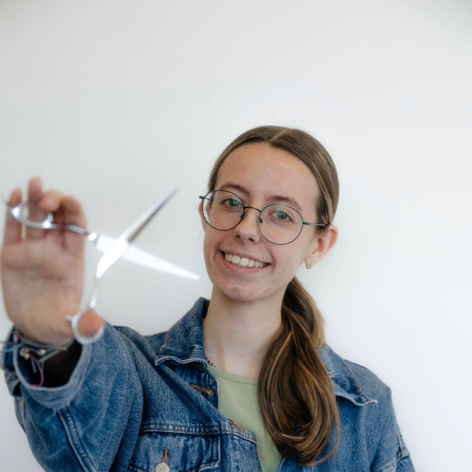 Woman holding scissors, smiling, wearing glasses and a black tank top, in sunny outdoor setting.