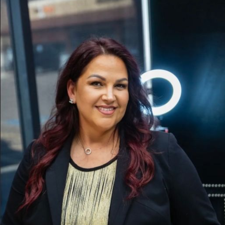 Woman with long wavy hair, in a black top, stands near a salon reception desk with mirrors and styling stations in the background.