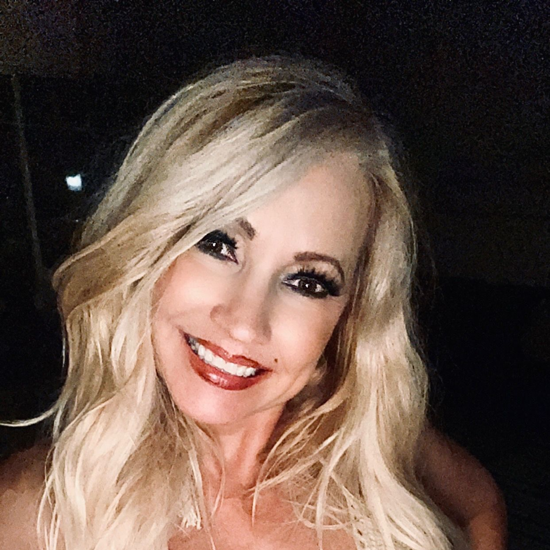 Blonde woman smiling, wearing lipstick, dark eye makeup, outdoors at night.