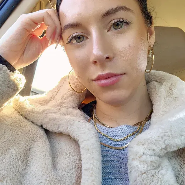 Woman with fair skin and light eyes wearing a cream-colored fuzzy jacket and gold jewelry.