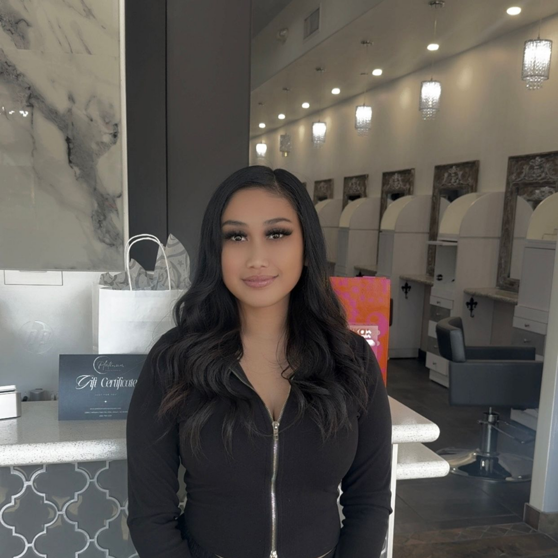 Woman with long wavy hair, in a black top, stands near a salon reception desk with mirrors and styling stations in the background.