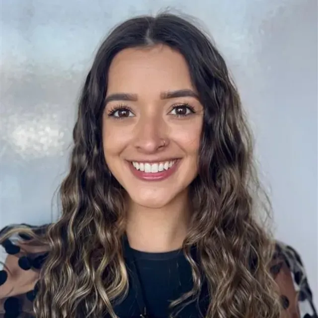 Woman with brown wavy hair smiles at the camera, wearing a black shirt with a polka-dot overlay.