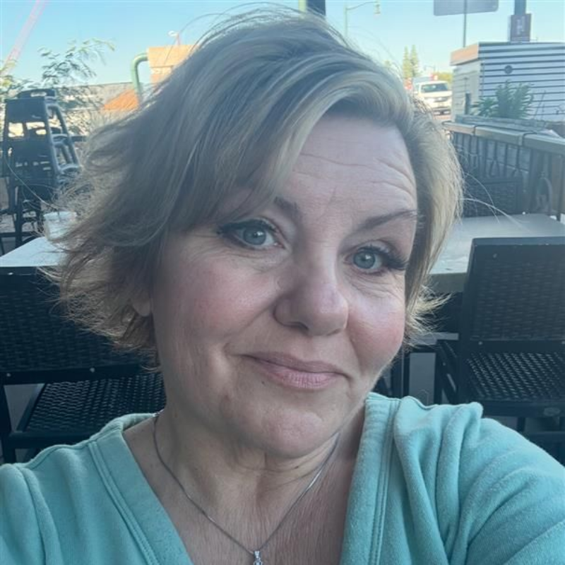 Woman with short blonde hair, smiling, in a teal shirt, seated outdoors at a table.