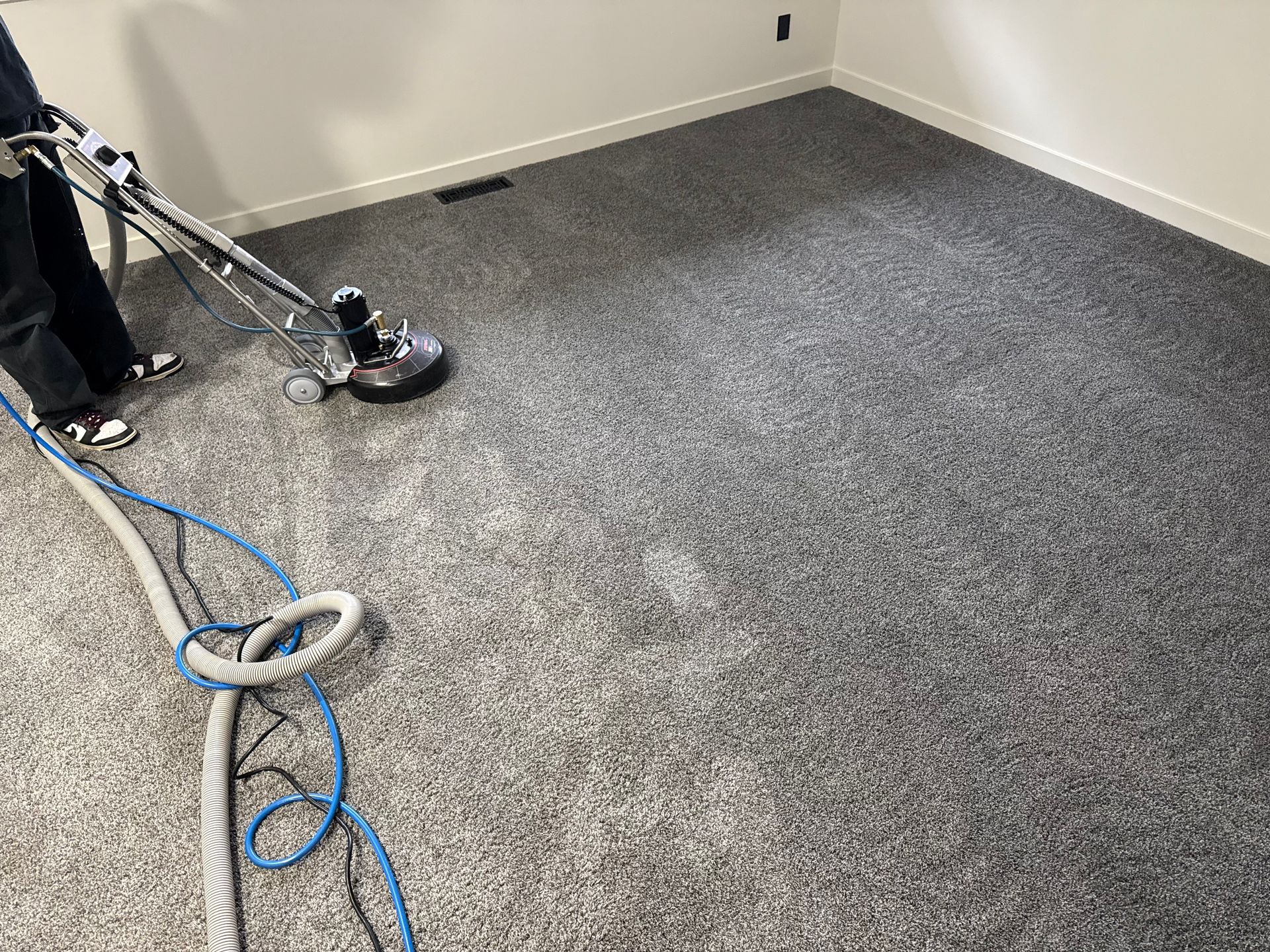 Person cleaning a gray carpet with a floor machine in a room with white walls.