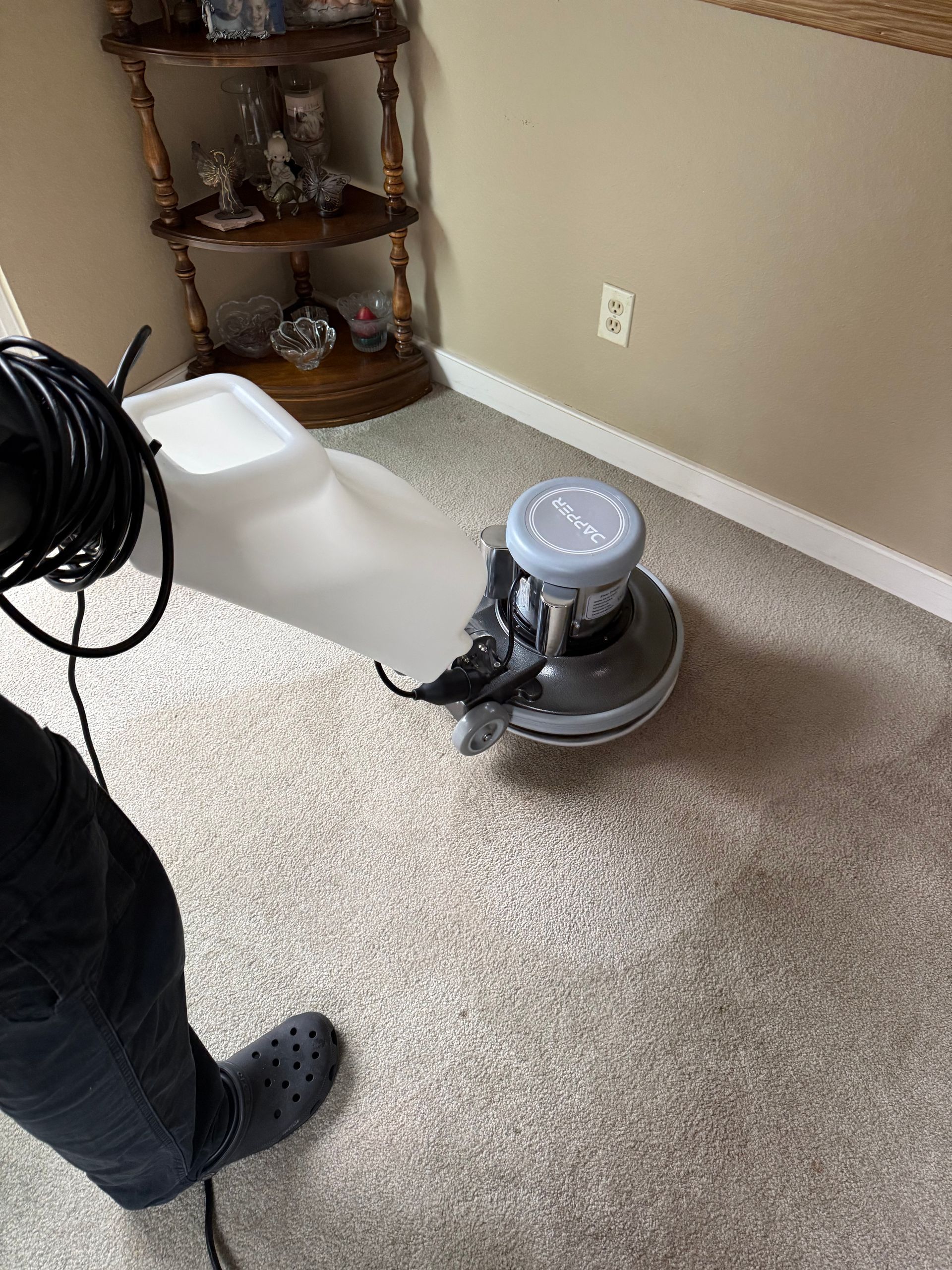 Person using a floor buffer on a carpeted floor, near a wall and furniture.