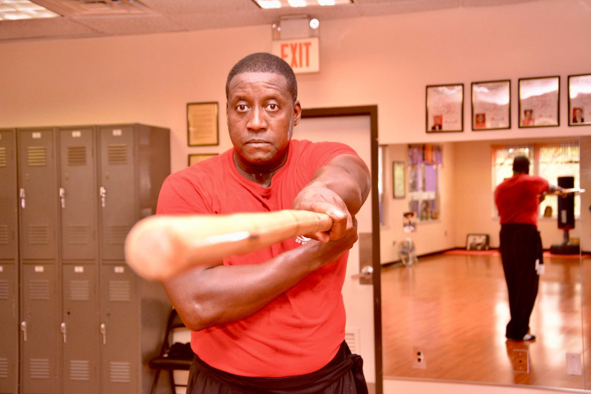 A man in a red shirt is holding a wooden bat in a gym.
