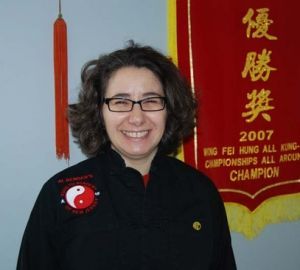 A woman wearing glasses is smiling in front of a banner that says 2007 king fei hung all kong championships all around champion