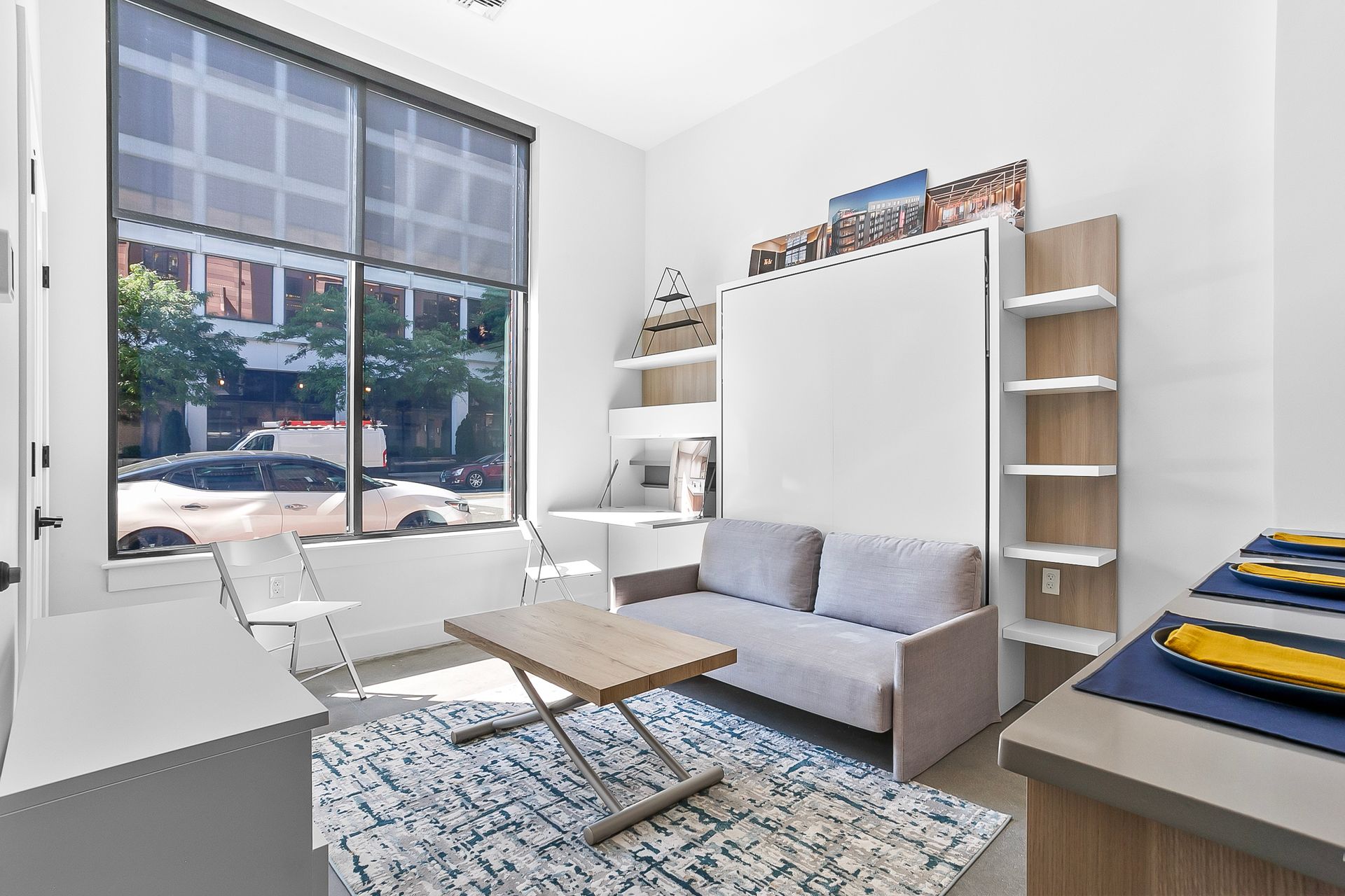 Small apartment with sofa, fold-out bed, shelves, rug, and large window overlooking a street.