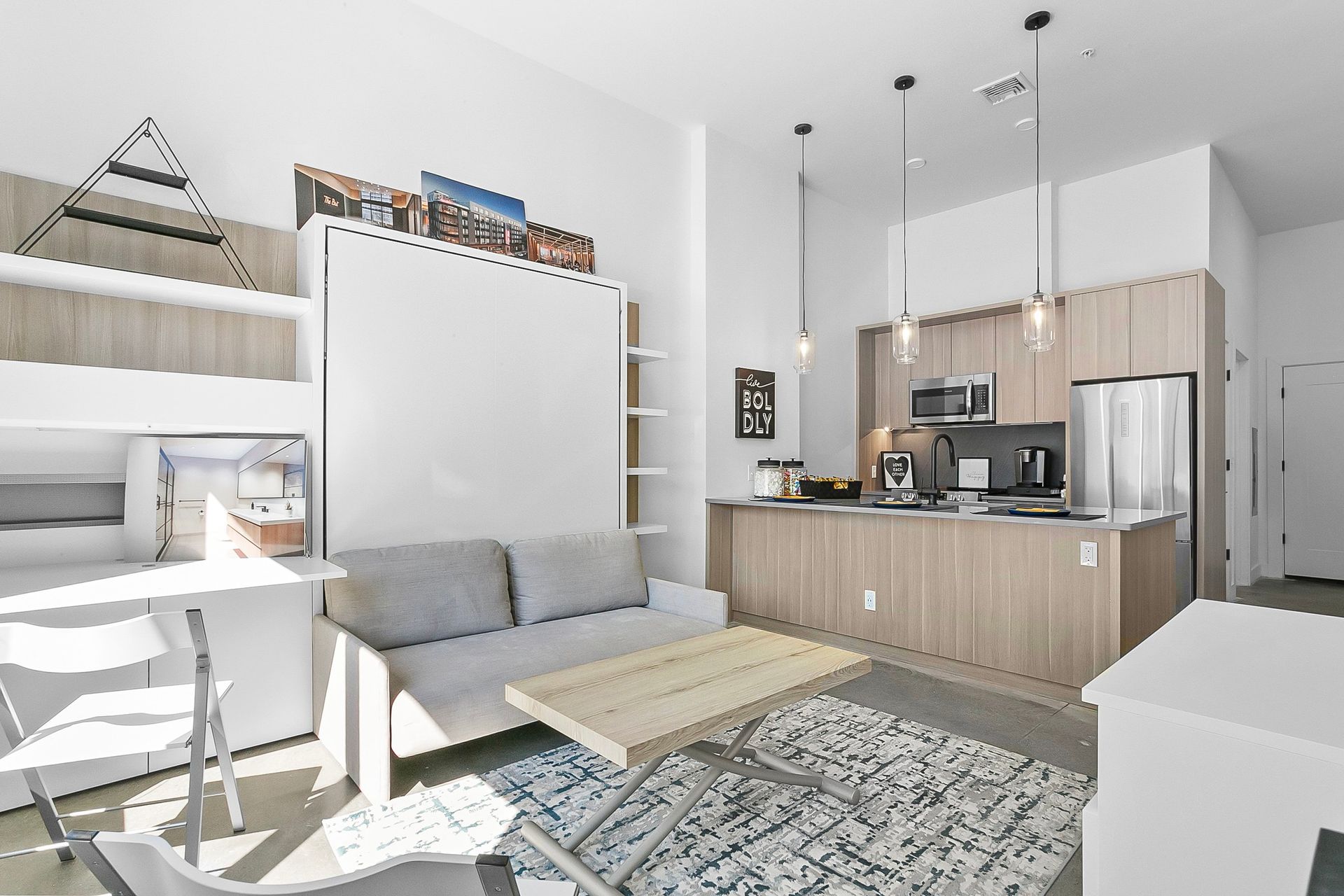 Small apartment interior with pull-down bed, kitchen, and living area. Natural wood cabinets, pendant lights.