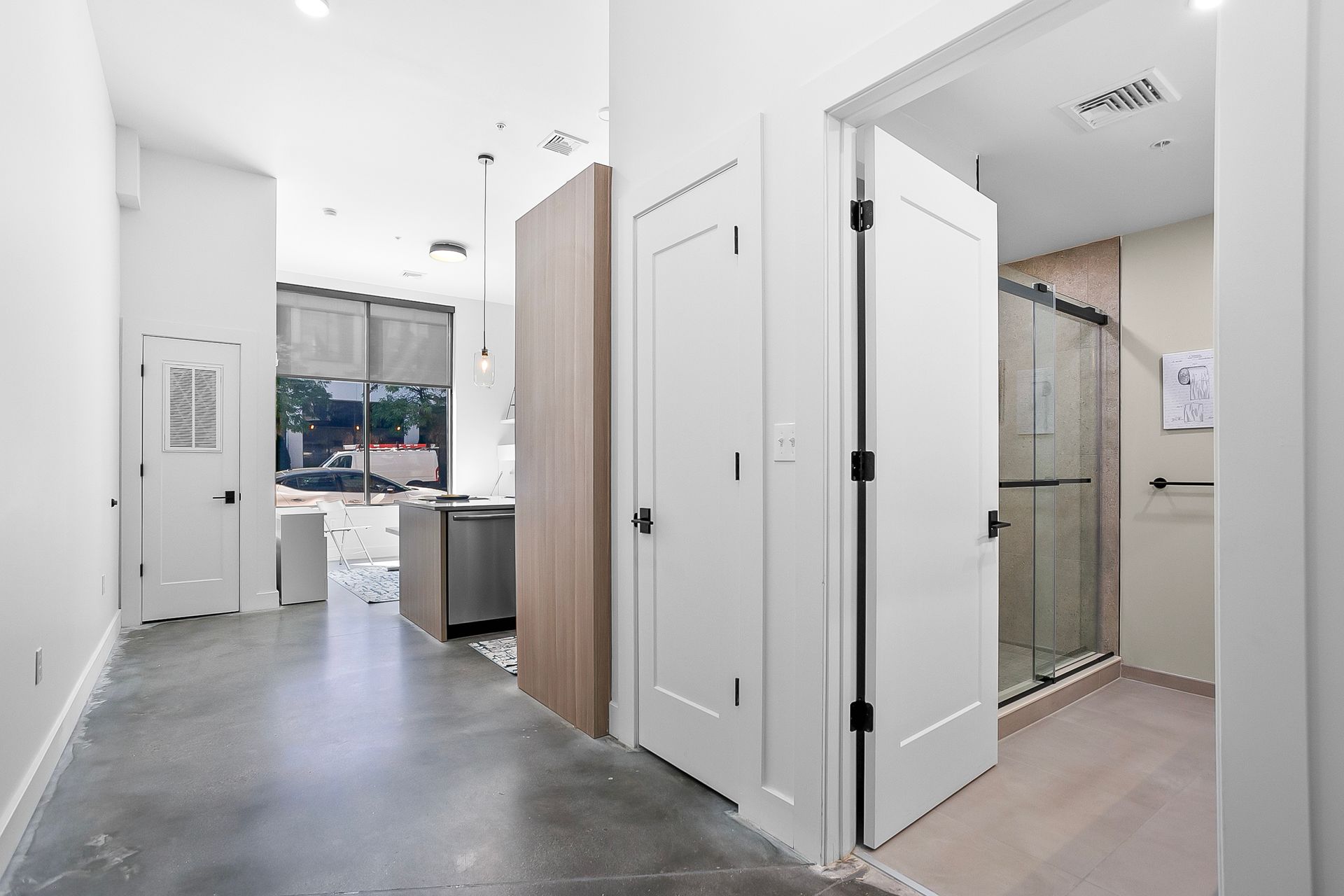 Interior view: hallway with white doors, open bathroom, and kitchen area with large windows.
