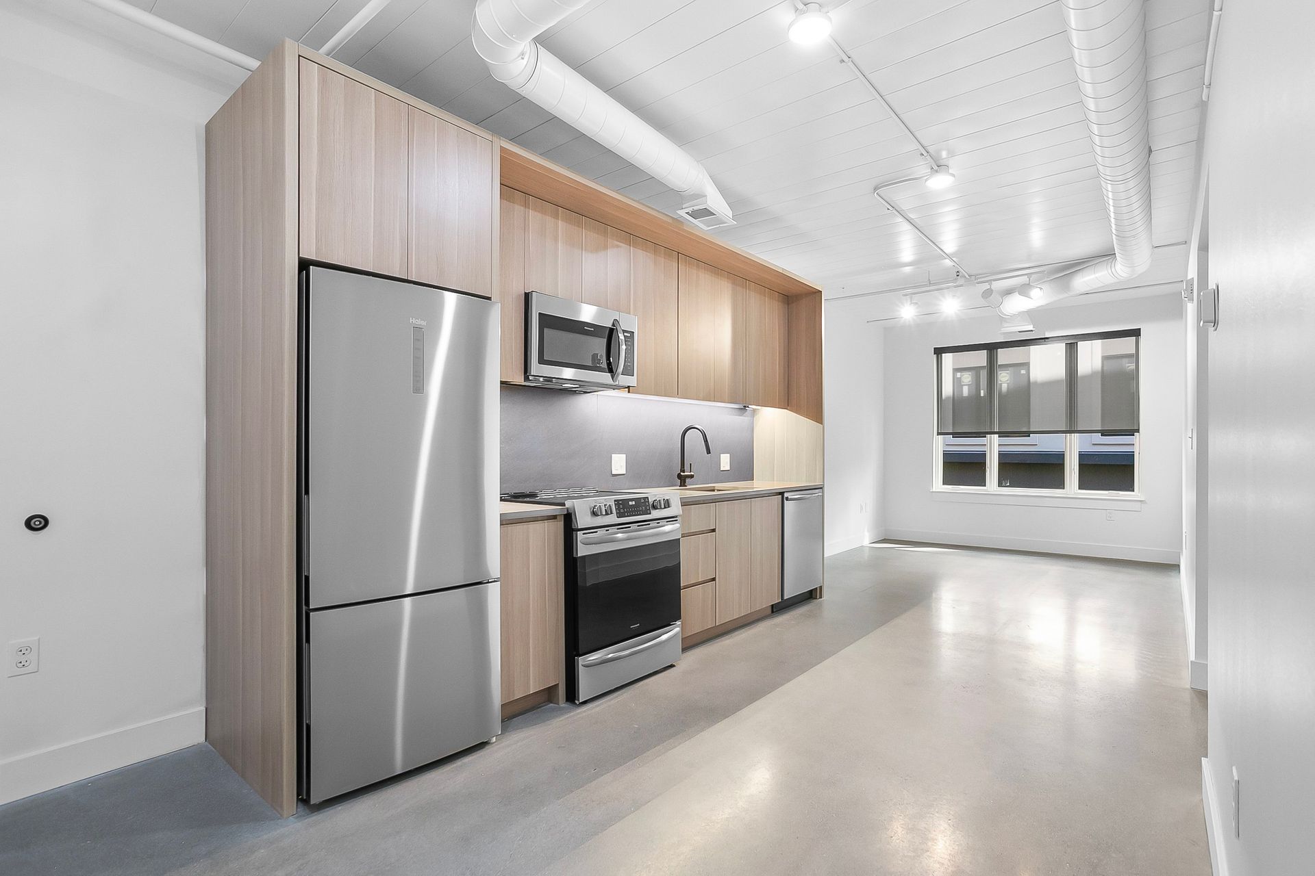 Modern apartment kitchen with stainless steel appliances, light wood cabinets, and concrete floor.