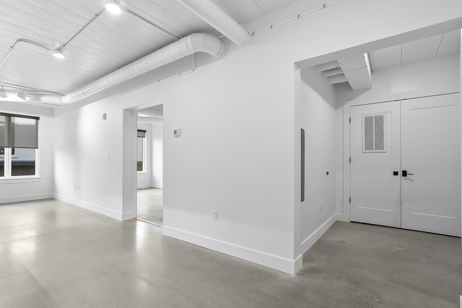 Empty, white-walled room with concrete floor, industrial ceiling fixtures, doorways, and a closed double door.