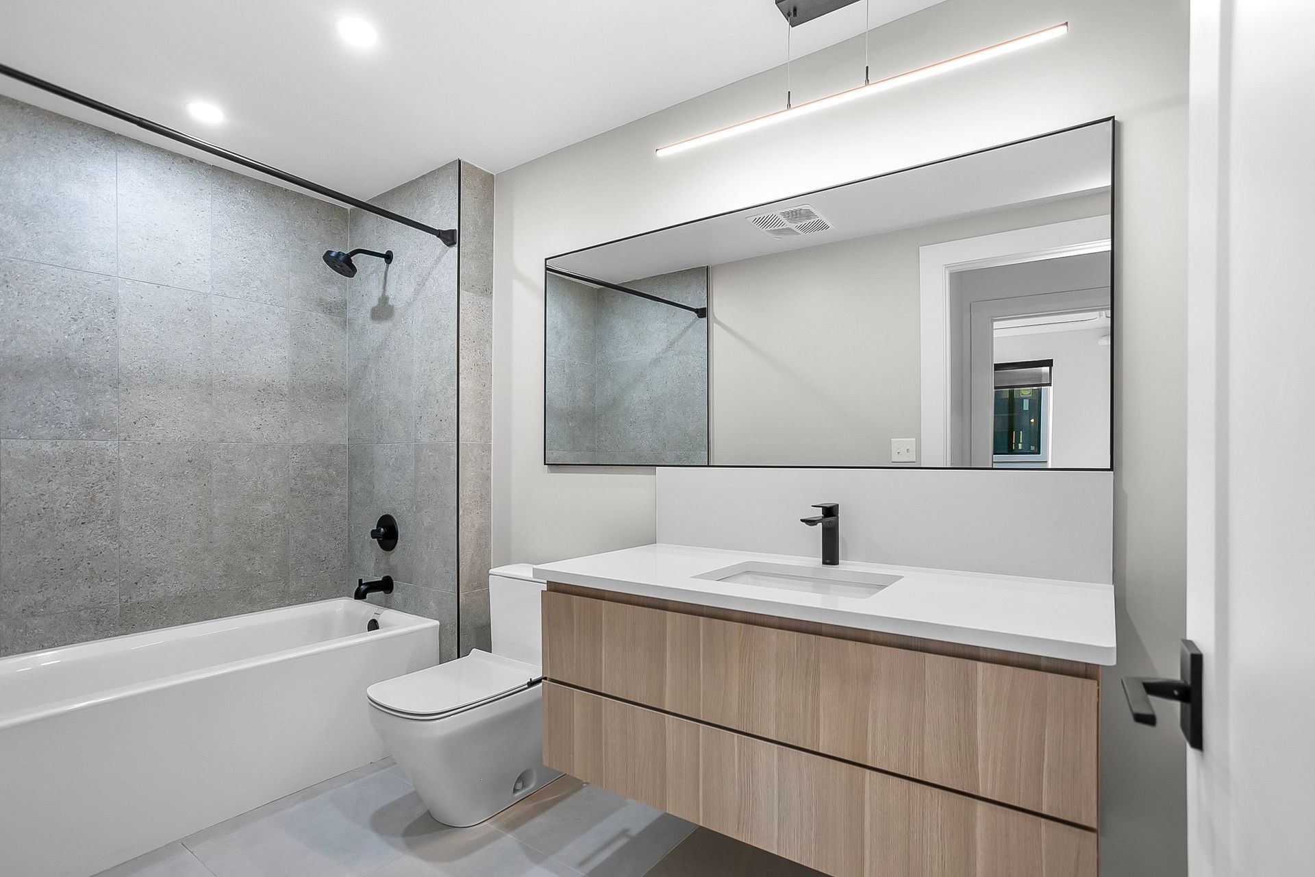 Modern bathroom with light wood vanity, white countertop, large mirror, gray tiled shower/tub, and black fixtures.