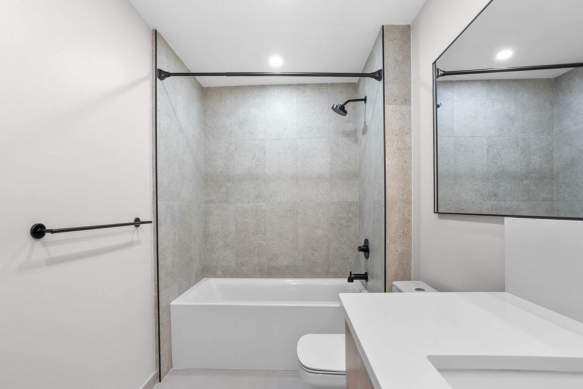 Bathroom with tub and shower, gray speckled tile and white sink. Black fixtures and a rectangular mirror.