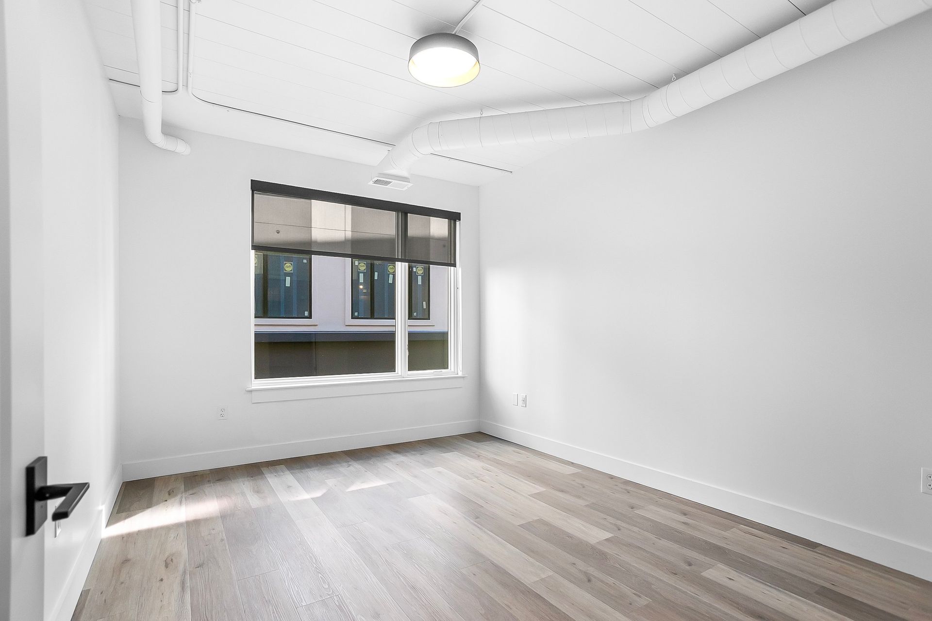 Empty, white-walled room with a window, hardwood floor, and a black door handle.