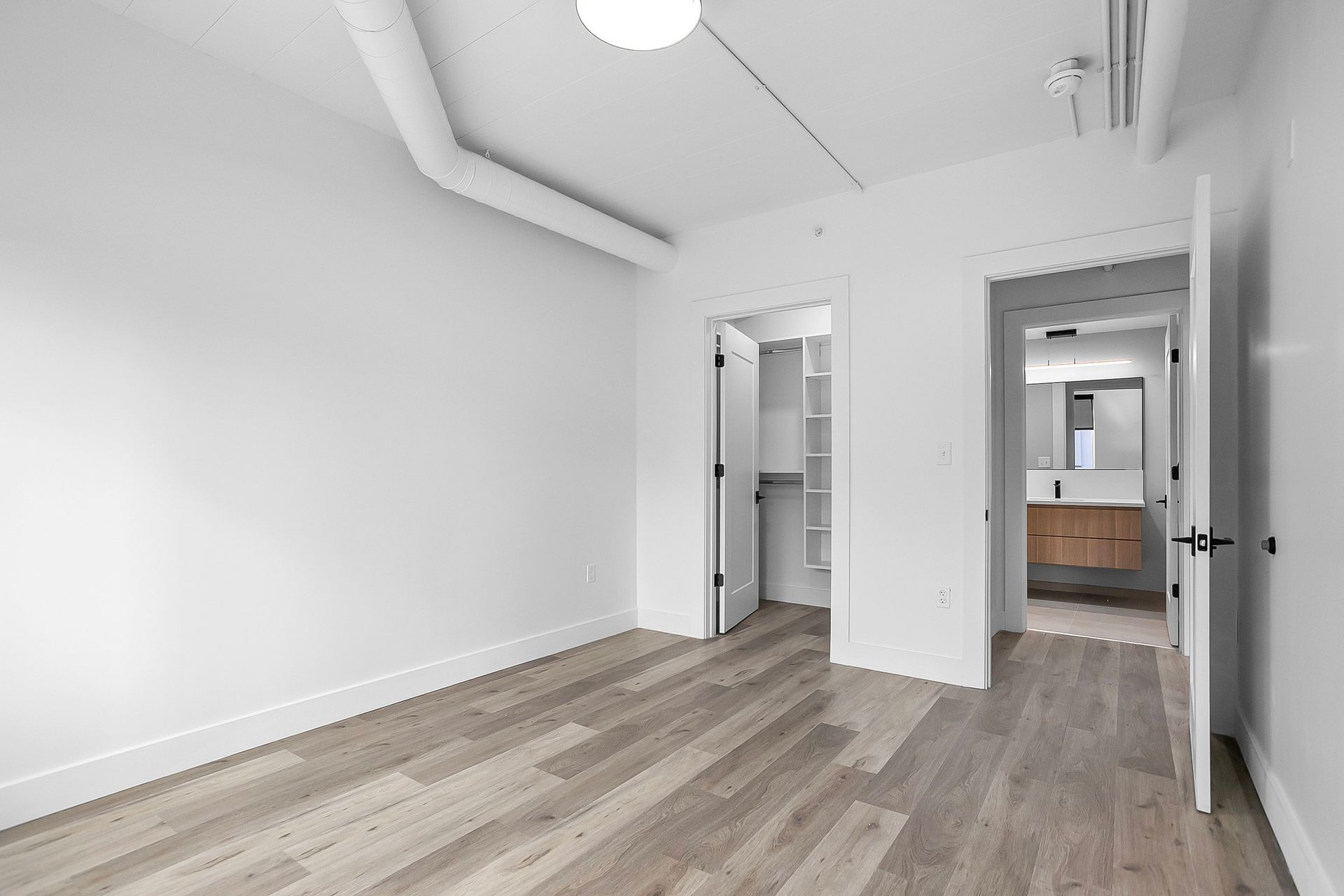 Empty, white bedroom with wooden floor, closet, and bathroom door open to a vanity.