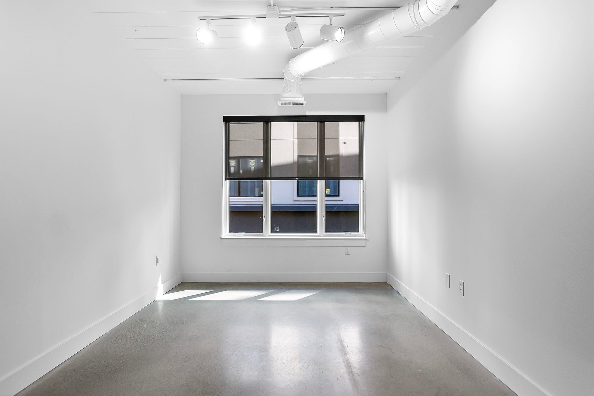 Empty, white-walled room with a large window, concrete floor, and ceiling fixtures.