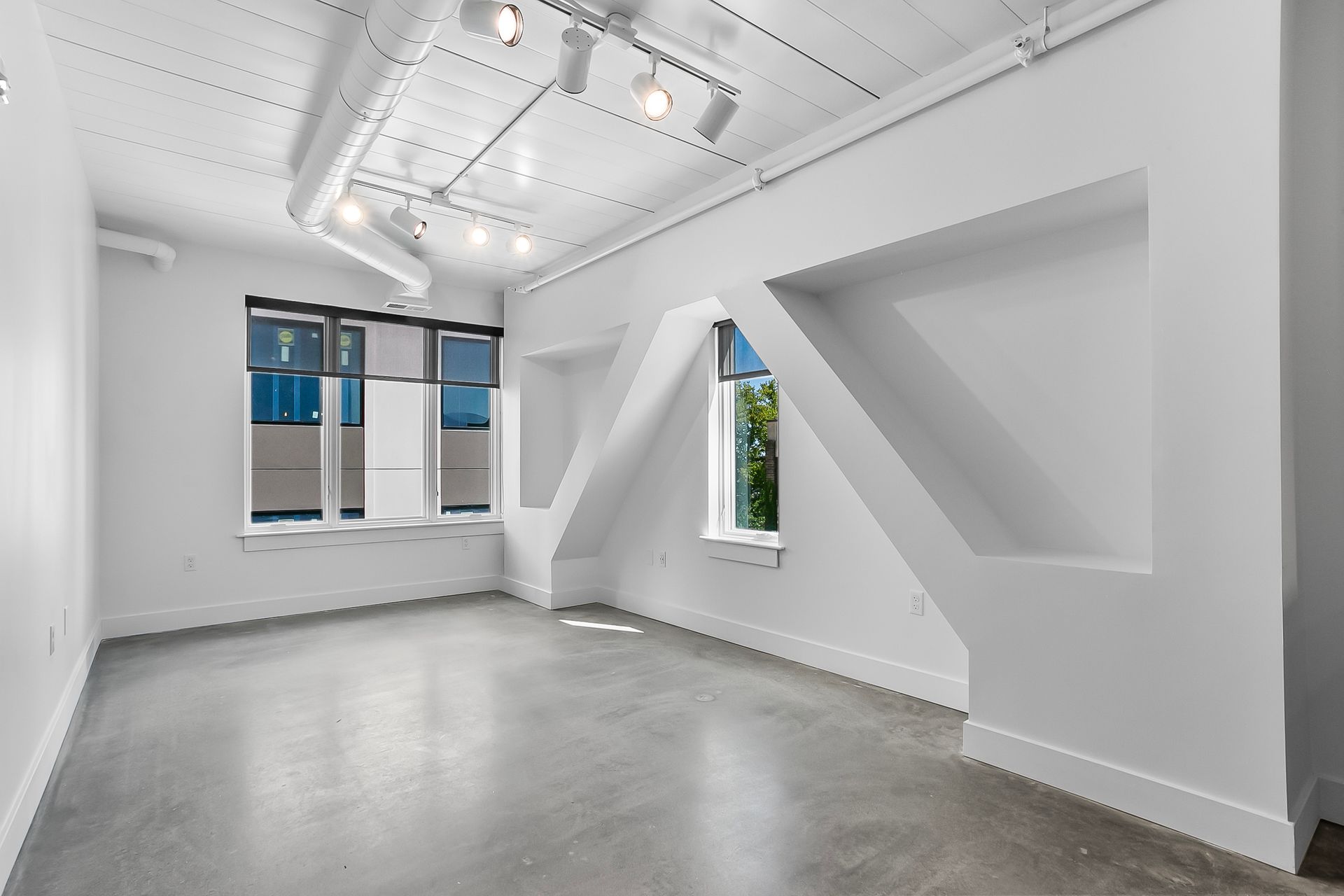 Empty, bright white room with large windows, concrete floor, and angled support beams.