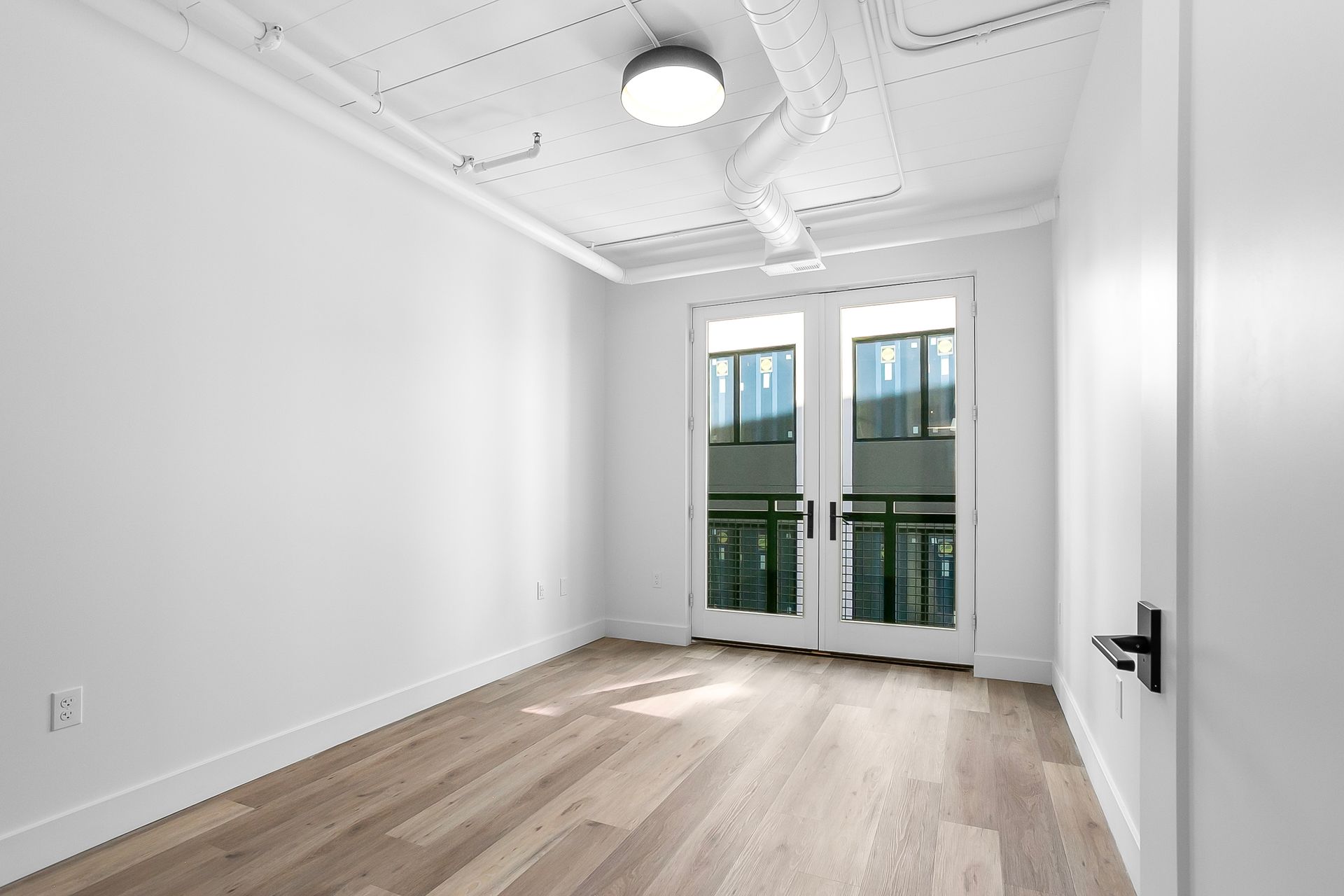 Empty room with wood floor, white walls, double doors to a balcony, ceiling light, and exposed pipes.